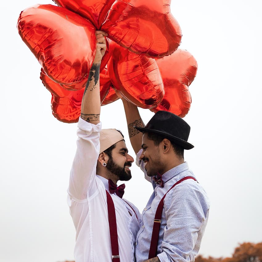 Gay couple smiles and holds up heart balloons on their wedding day