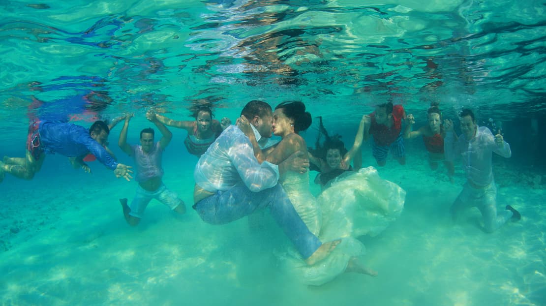  Couple kissing underwater while surrounded by friends 